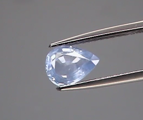 Pear-shaped blue gemstone held by tweezers against a gray background