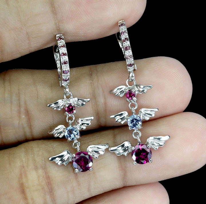 Silver angel wing earrings with gemstones held between fingers against a dark background