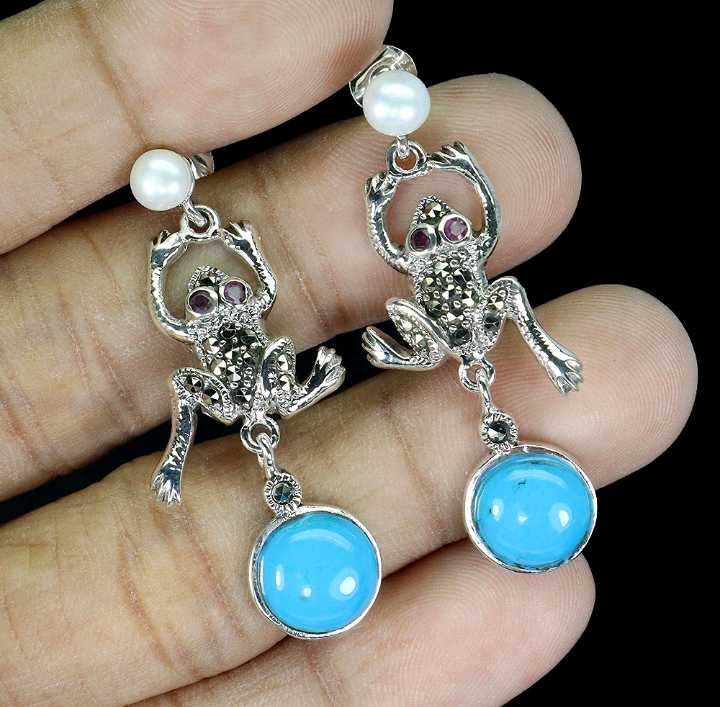 Frog-shaped earrings with blue stones and pearl accents held between fingers against a black background