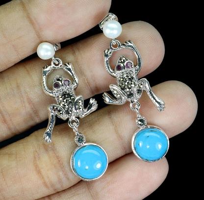 Frog-shaped earrings with blue stones and pearl accents held between fingers against a black background